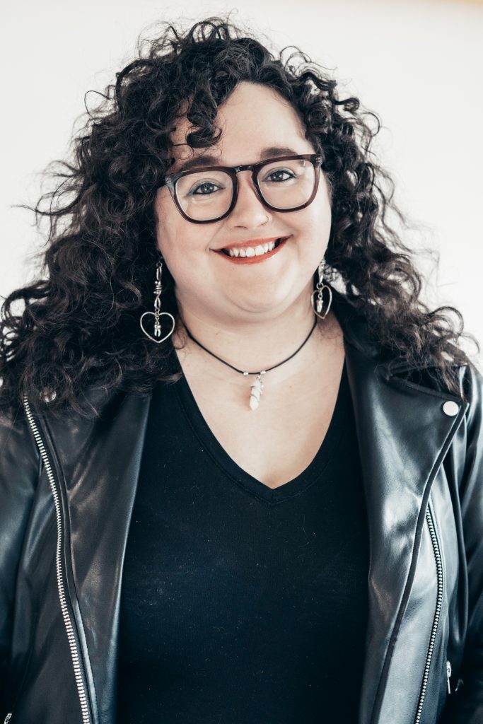 Portrait photo of myself smiling, a white woman with brown shoulder length curly hair. I am wearing a black leather biker jacket and eyeglasses.