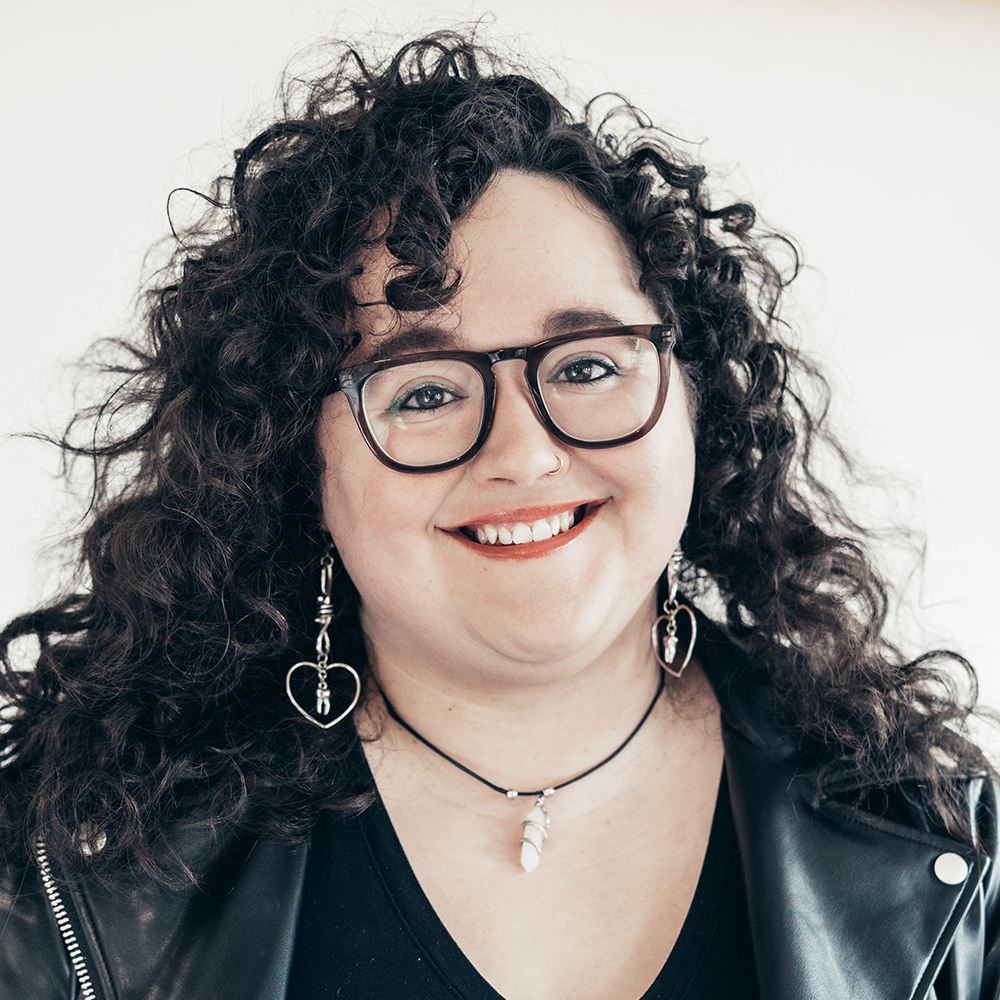 Headshot of myself smiling, a white woman with shoulder-length brown curly hair. I wear eyeglasses.